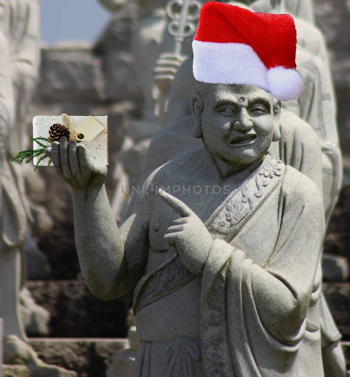 Christmas stone buddhist monk statue pointing at the gift with blank card he is holding and wearing a red santa claus hat by charlottebleijenberg