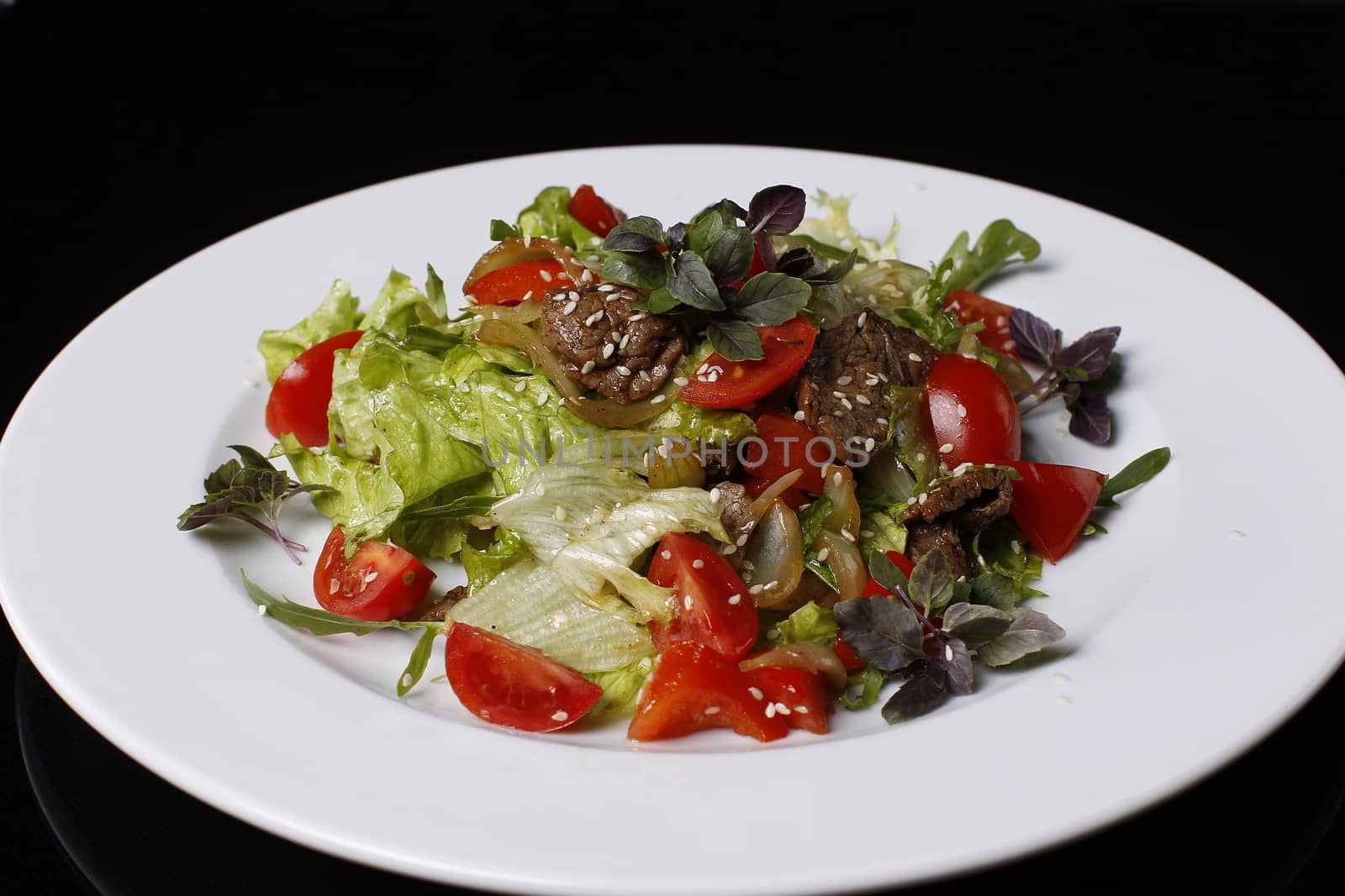 On a white plate salad of tomatoes, fried meat, onions, lettuce, cabbage strewed with sesame seeds, basil, on a black background.Dietary food by Sviatlana