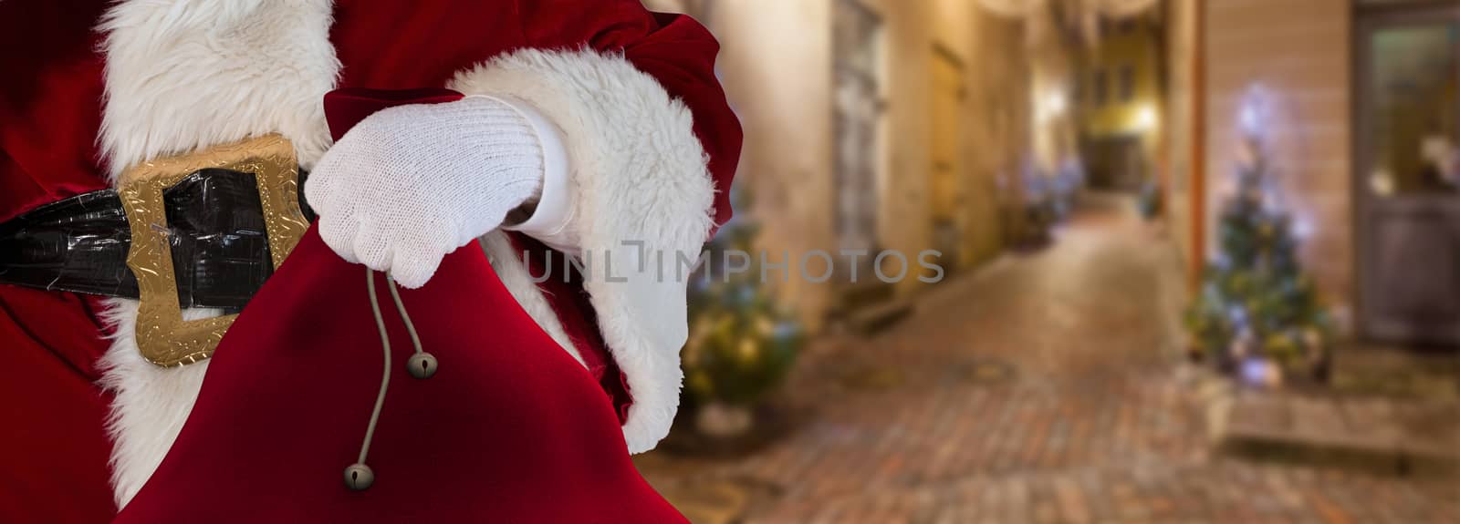 Christmas concept Santa claus standing on the streets of a small alley holding his bag full of presents by charlottebleijenberg