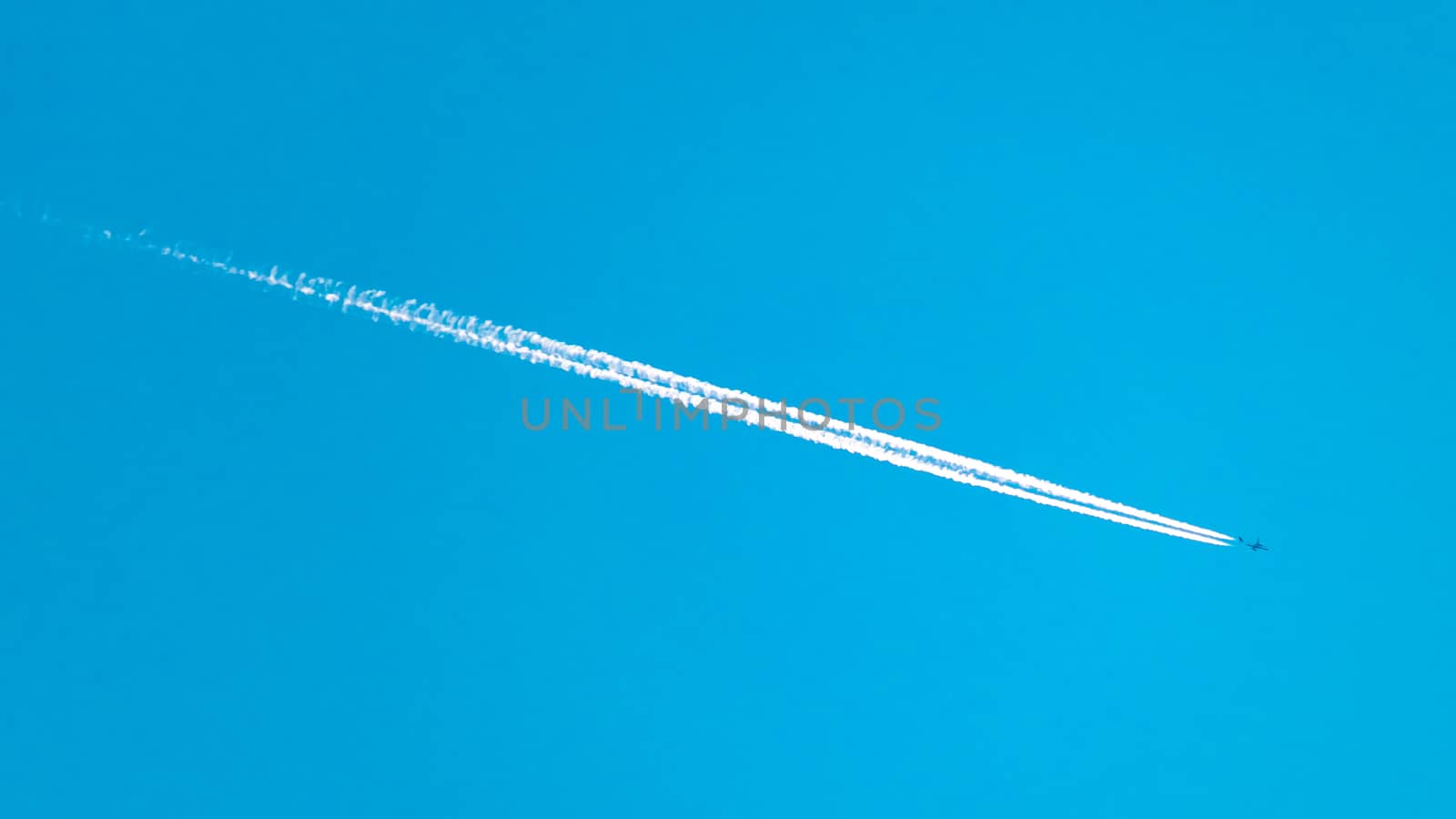 An airplane flies high overhead, leaving a white vapor trail streaking across the blue sky behind it.