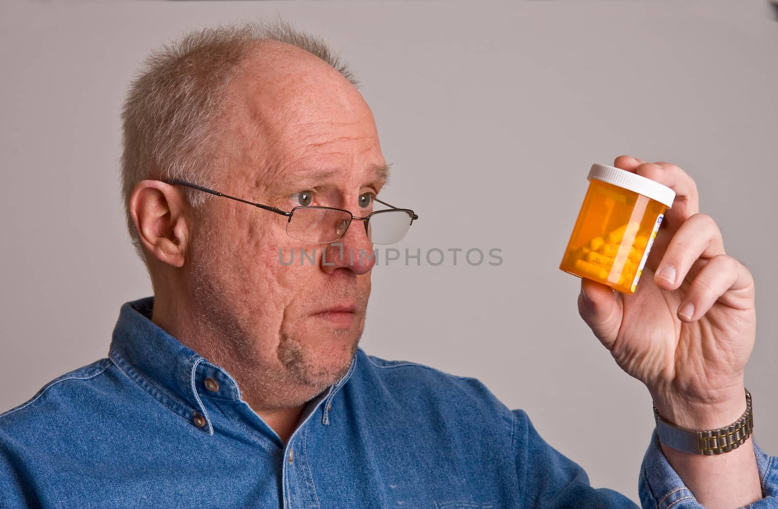 An older bald man in a blue denim shirt on a grey background wearing glasses and looking at a prescription bottle