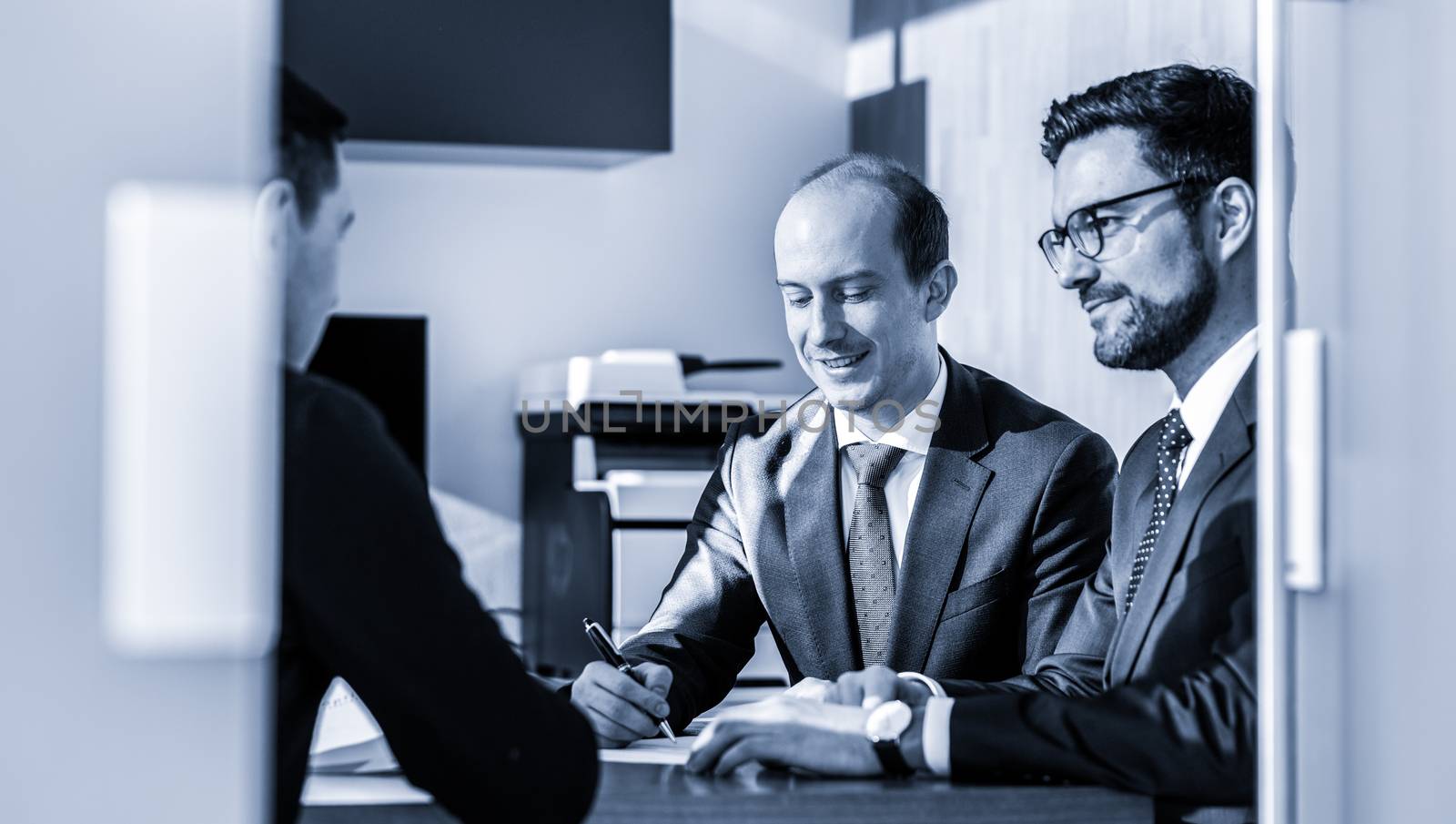 Team of confident successful business people reviewing and signing a contract to seal the deal at business meeting in modern corporate office. Business concept. Greyscale blue toned image.