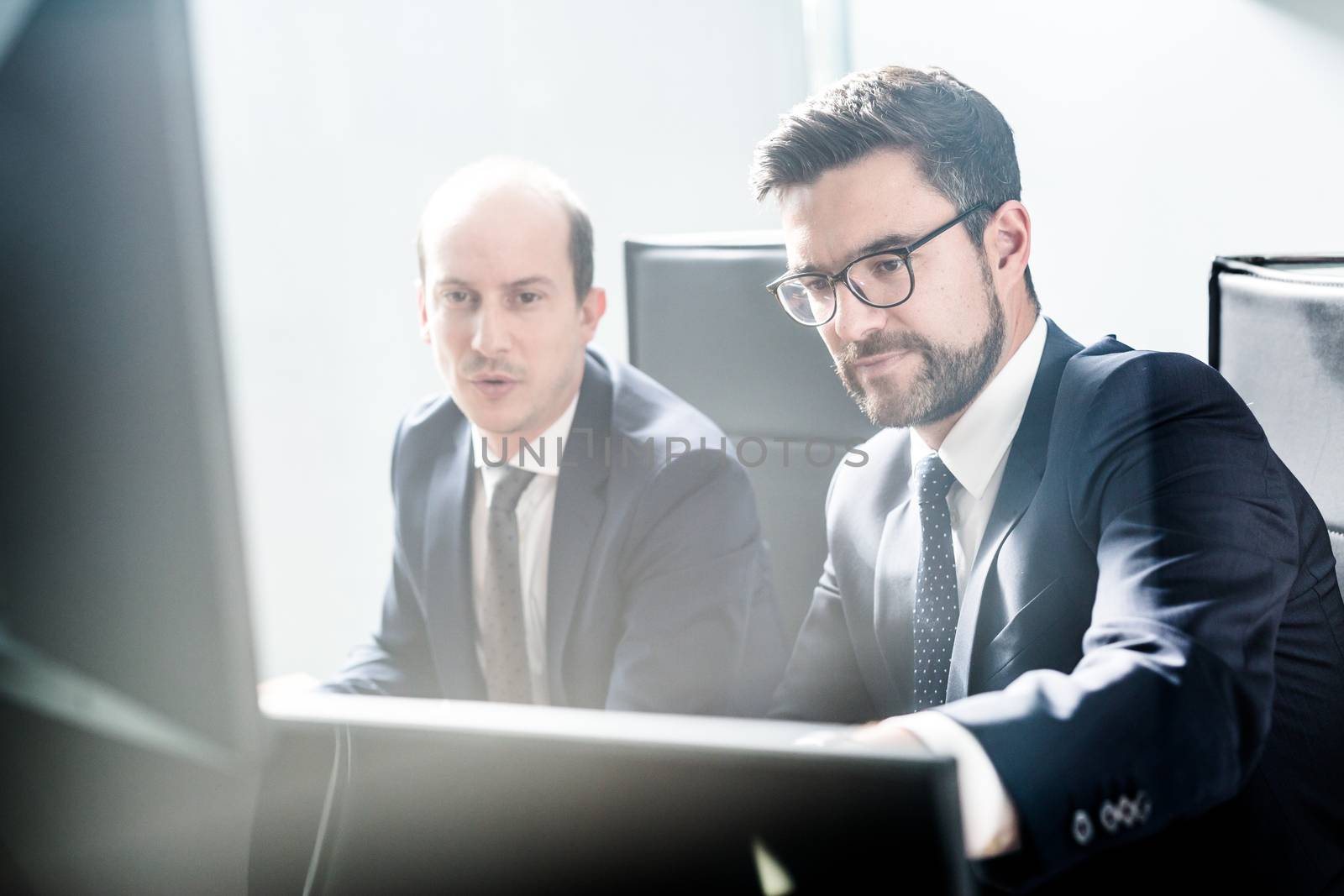 Image of two thoughtful businessmen looking at data on multiple computer screens, solving business issue at business meeting in modern corporate office. Business success concept.