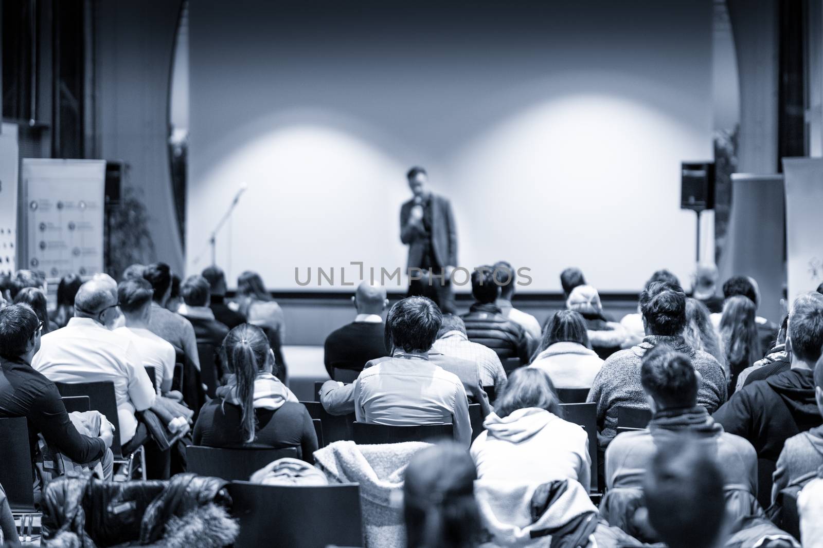 Male public speaker giving a talk at business meeting conference. Rear view of unrecognized people sitting in audience in the conference hall. Business and entrepreneurship concept. Blue toned.