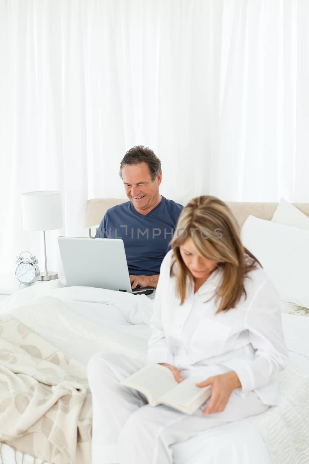 Senior looking at his laptop while her wife is reading at home