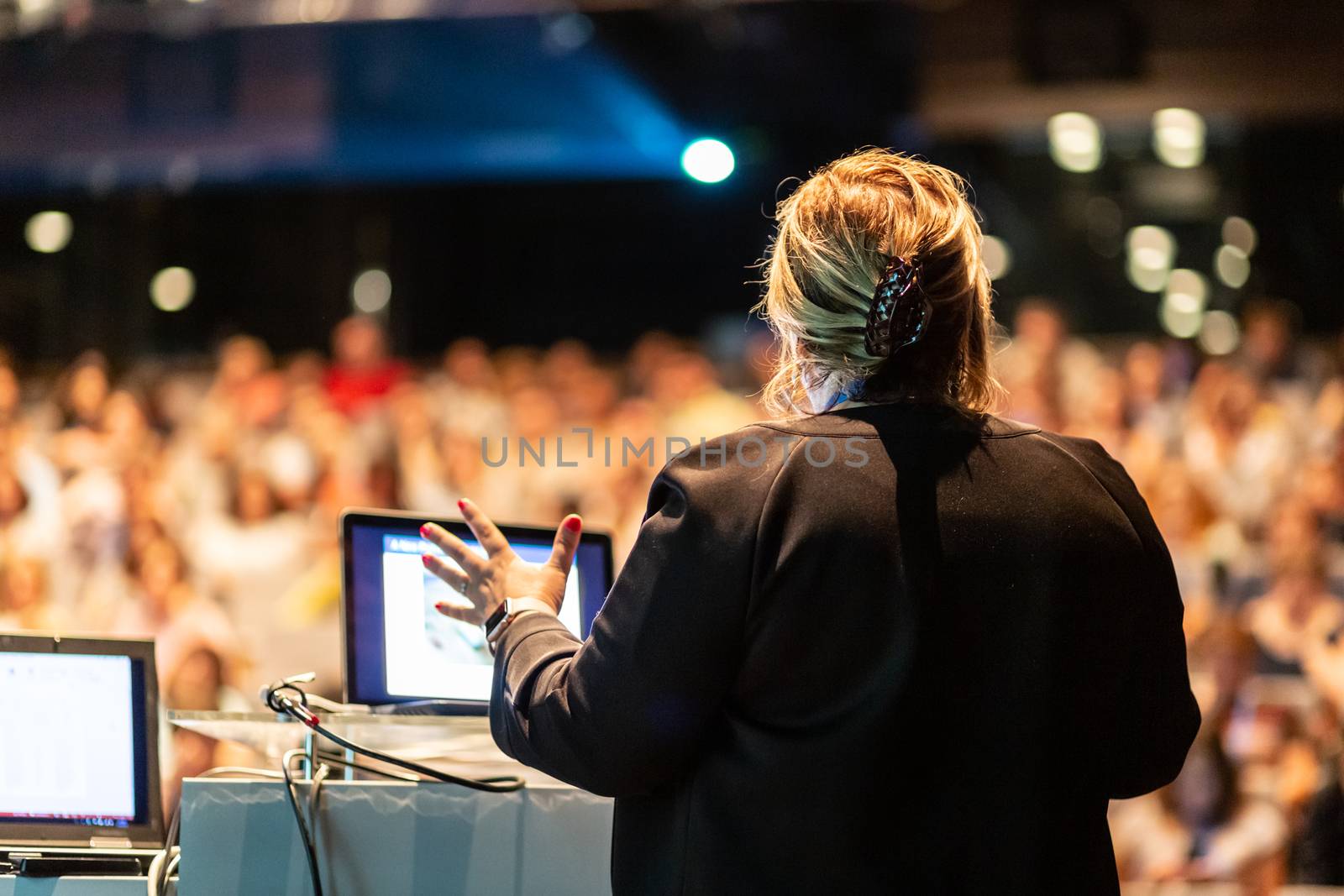 Female speaker giving a talk on corporate business conference. Unrecognizable people in audience at conference hall. Business and Entrepreneurship event.