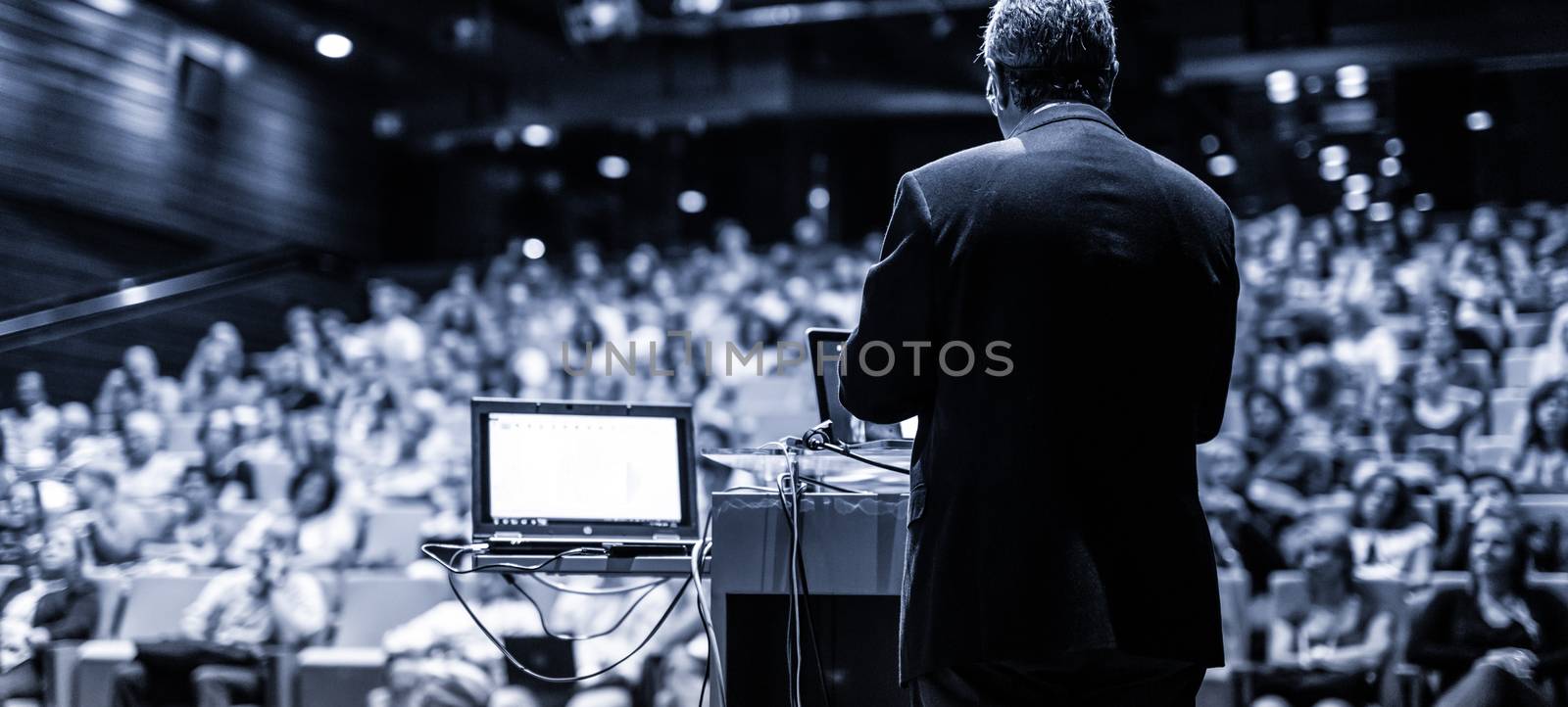 Speaker giving a talk on corporate business conference. Unrecognizable people in audience at conference hall. Business and Entrepreneurship event. Black and white, blue toned image.