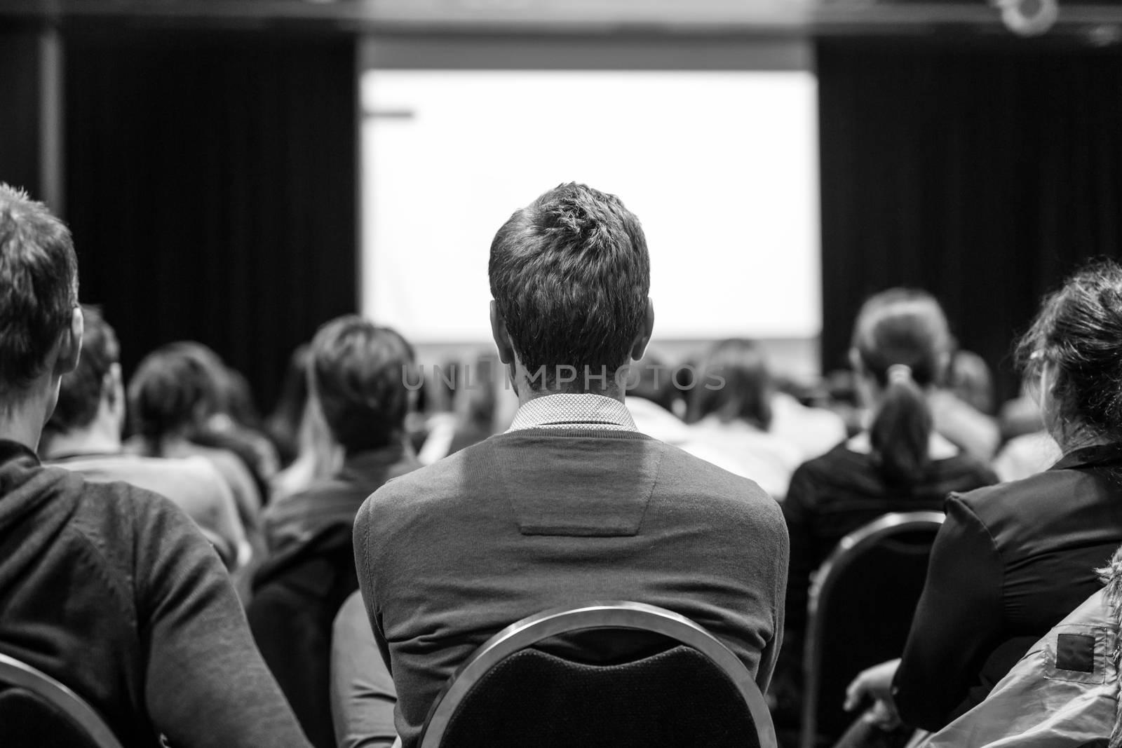 Business and entrepreneurship symposium. Speaker giving a talk at business meeting. Audience in conference hall. Rear view of unrecognized participant in audience. Black and white.