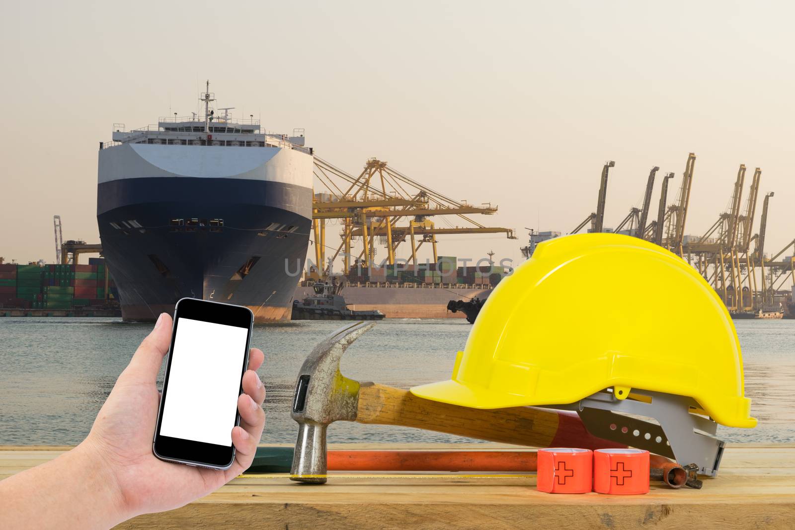 Hand holding the black smartphone with Yellow Safety Helmet on foreground of 
huge container cargo ship at harbor.
