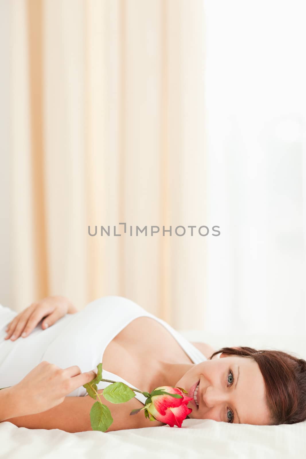 Woman with a rose lying on a bed looking into camera in her bedroom