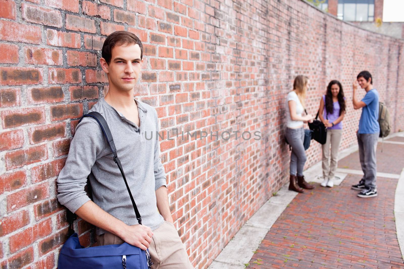 Student leaning on a wall while his friends are talking outside a building