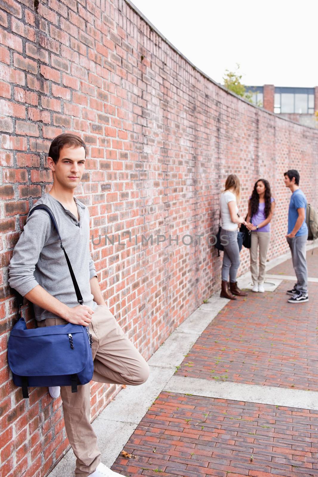 Portrait of a student leaning on a wall while his friends are talking outside a building