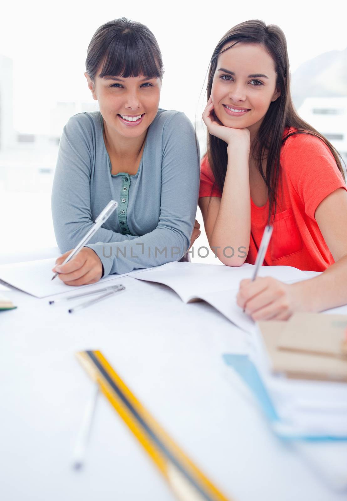 A pair of smiling girls looking at the camera as they work on schoolwork by Wavebreakmedia
