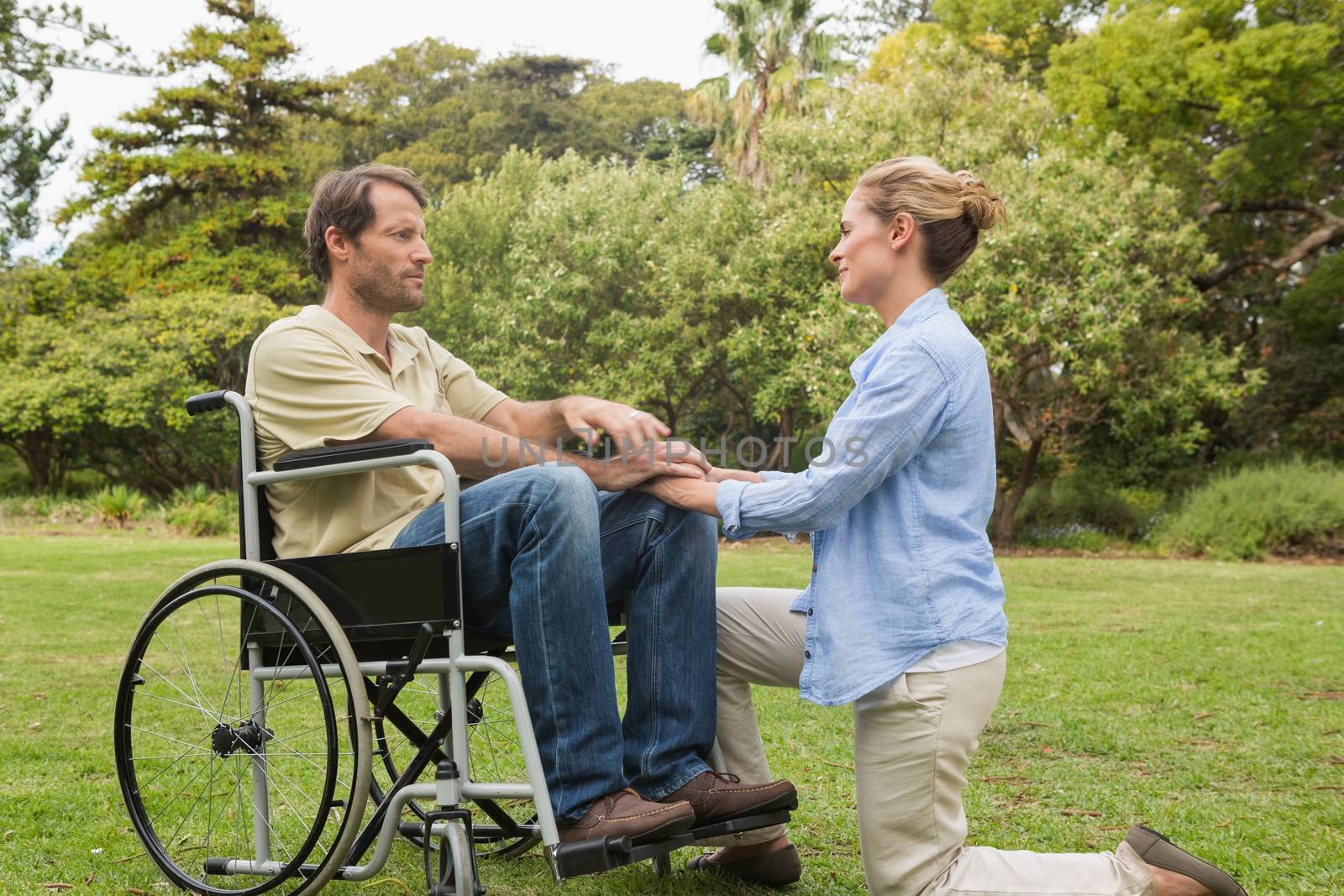 Content man in wheelchair with partner kneeling beside him and looking at each other in a park