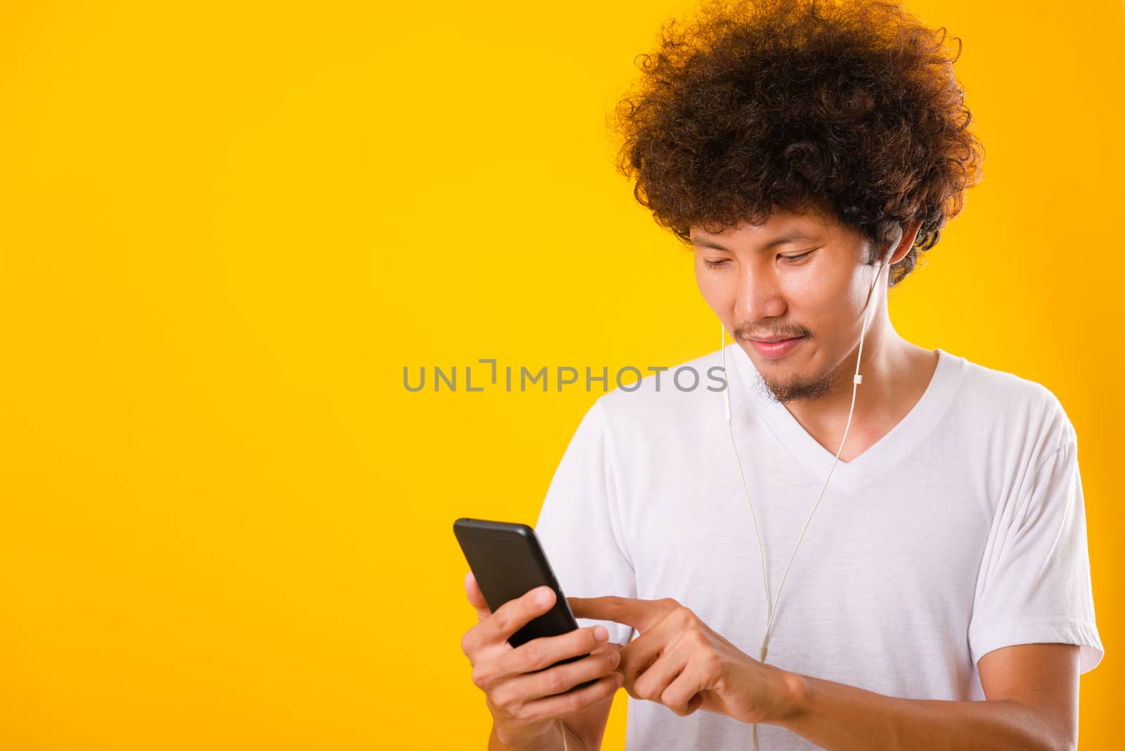 Happy asian handsome man with curly hair he smiling enjoying listening to music on earphones using a mobile smartphone isolate on yellow background