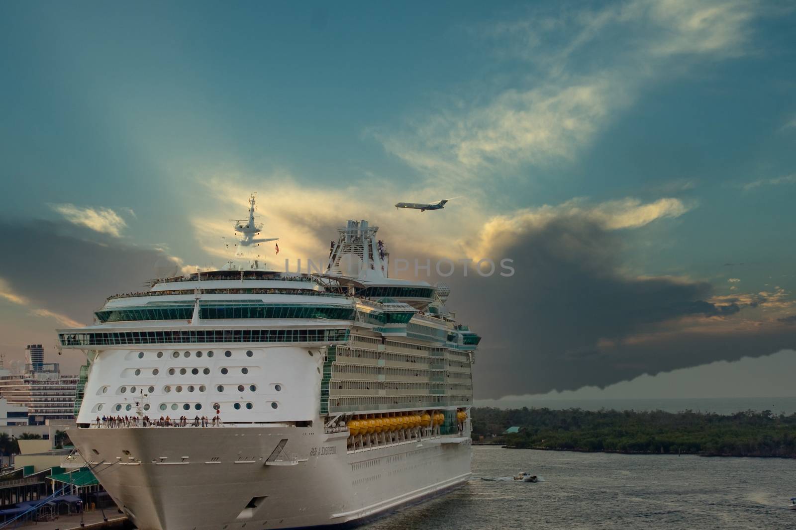 A large cruise ship anchored at a pier with a commercial airplane flying over at dusk