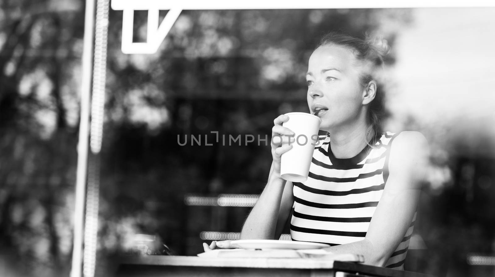 Young caucasian woman sitting alone in coffee shop drinking american coffee, people watching, thoughtfully looking trough the coffee shop window. Black and white image.