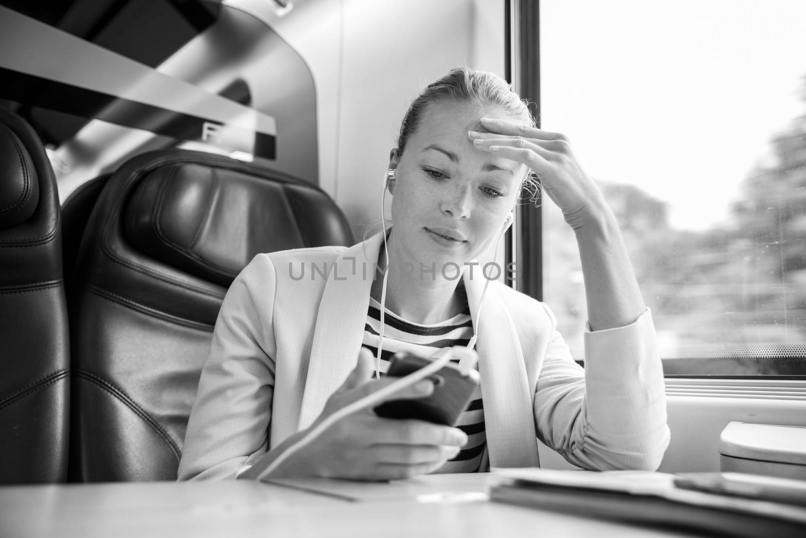 Caucasian businesswoman communicating on cellphone using headphone set while traveling by train in business class seat. Black and white image.