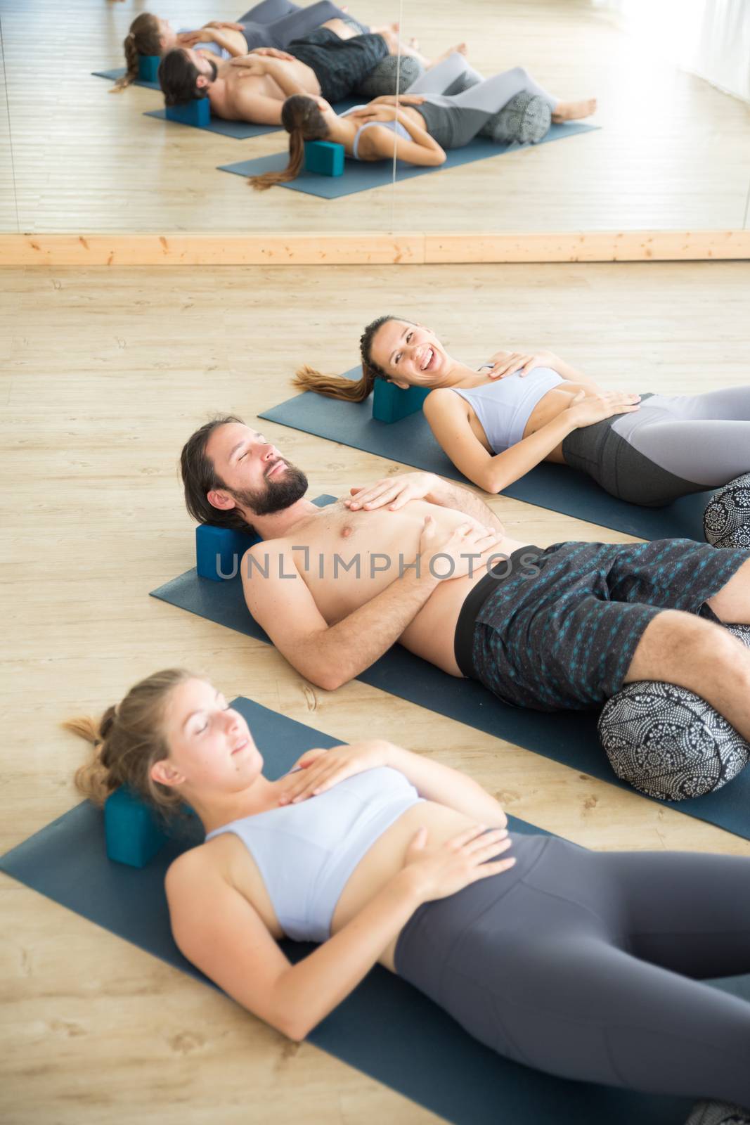 Restorative yoga with a bolster. Group of young sporty attractive people in yoga studio, lying on bolster cushion, stretching and relaxing during restorative yoga. Healthy active lifestyle.