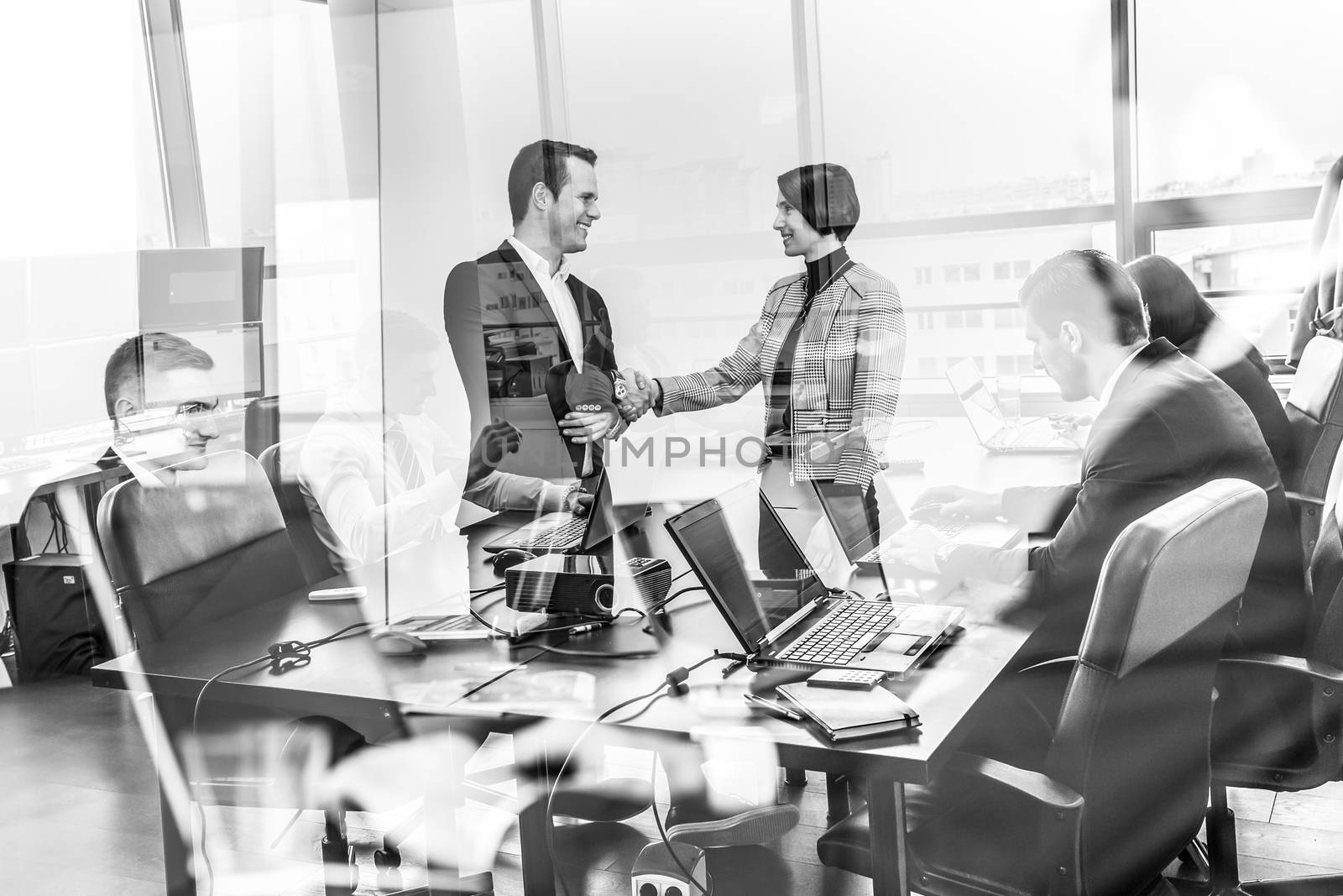 Sealing a deal. Business people shaking hands, finishing up meeting in corporate office. Businessmen working on laptops seen in glass reflection. Business concept in black and white.
