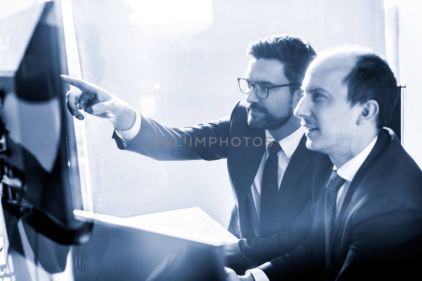Image of two thoughtful businessmen looking at data on multiple computer screens, solving business issue at business meeting in modern corporate office. Business success concept. Blue toned.