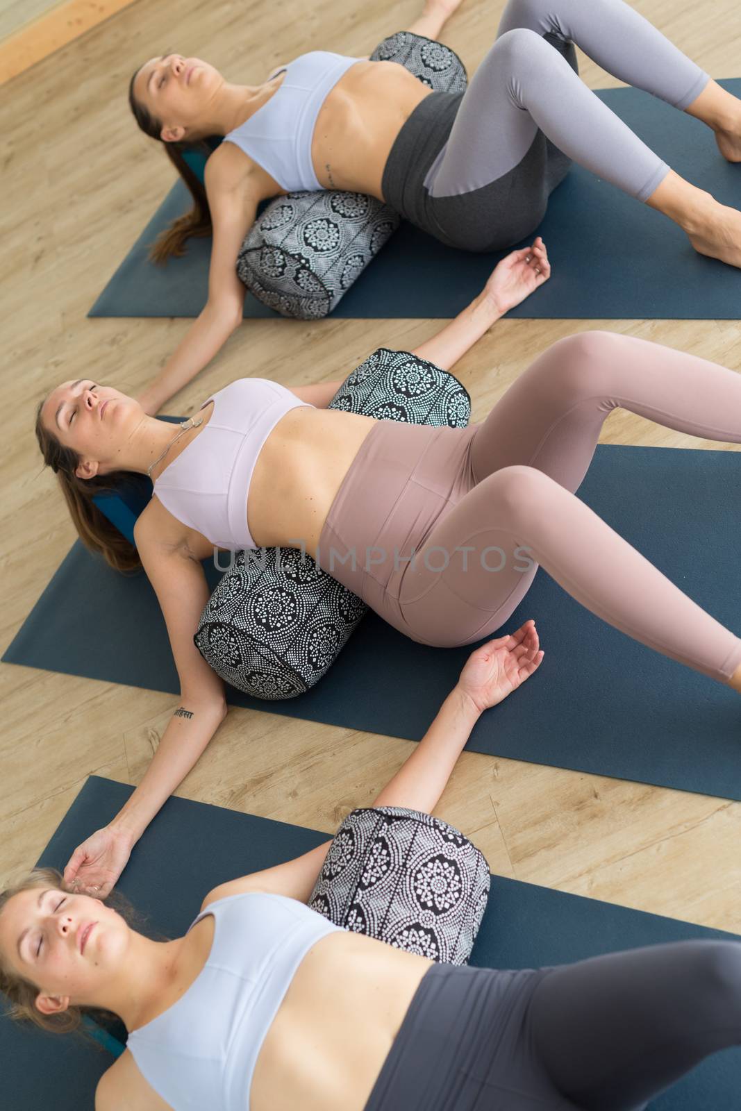 Restorative yoga with a bolster. Group of three young sporty attractive women in yoga studio, lying on bolster cushion, stretching and relaxing during restorative yoga. Healthy active lifestyle.
