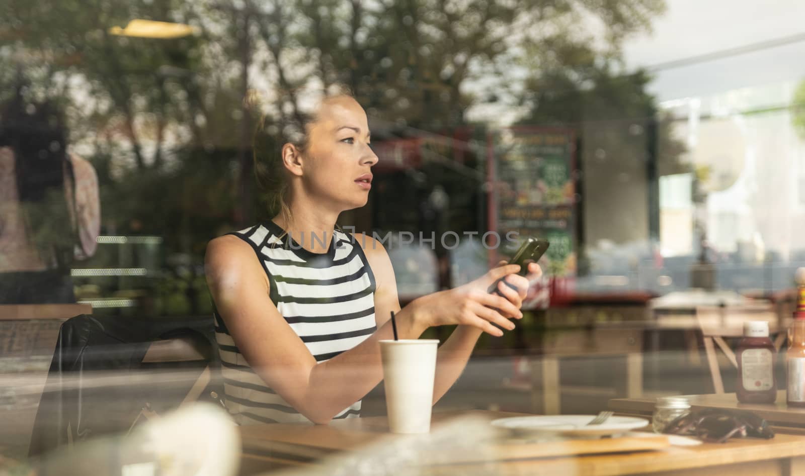 Thoughtful caucasian woman holding mobile phone while looking through the coffee shop window during coffee break. by kasto