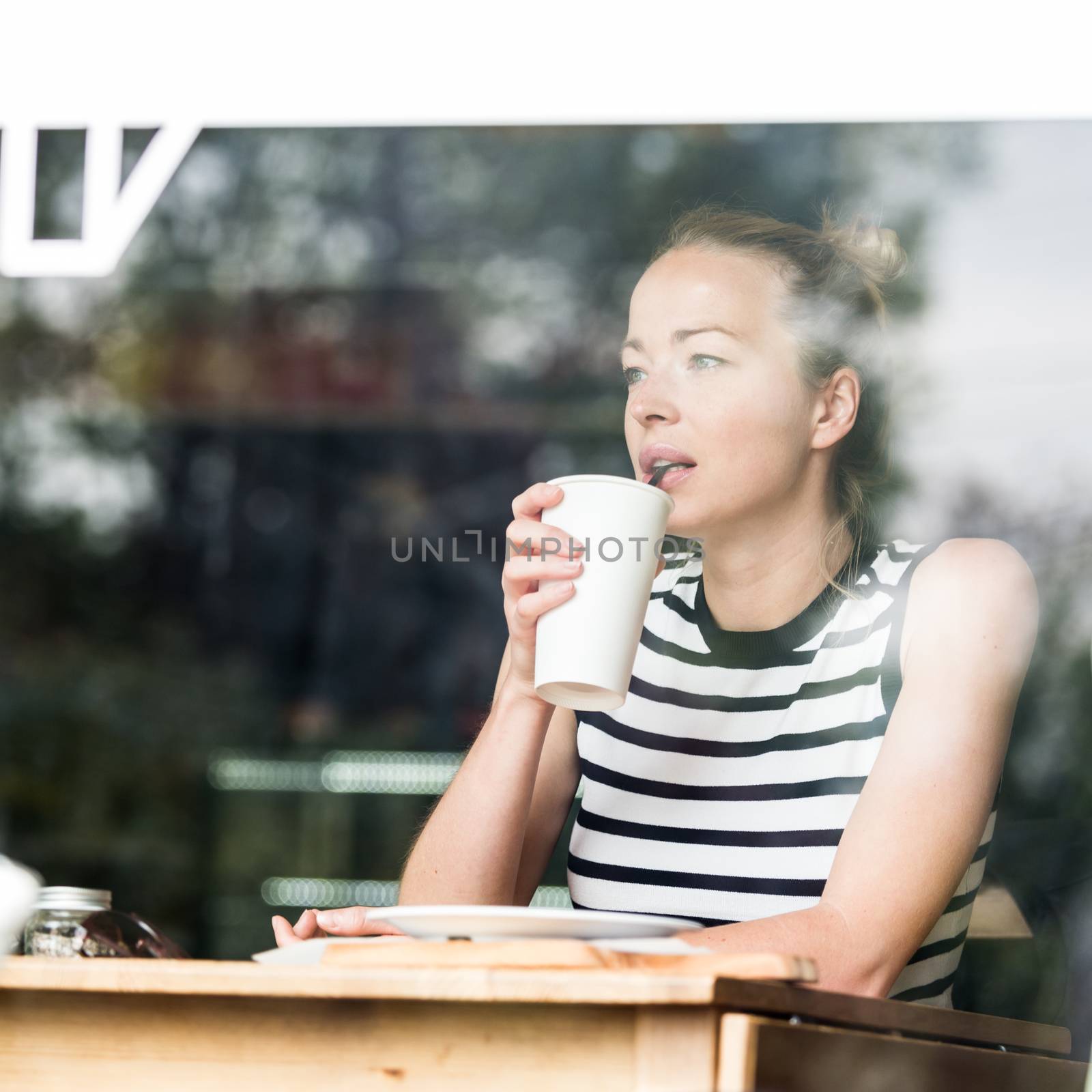 Young caucasian woman sitting alone in coffee shop drinking american coffee, people watching, thoughtfully looking trough the coffee shop window.