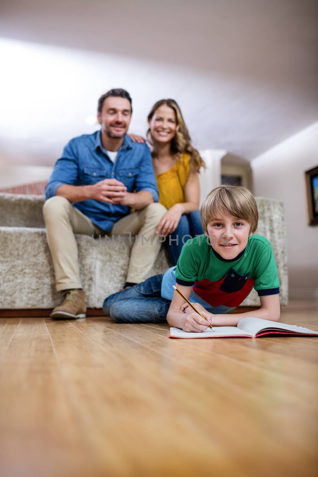 Boy doing homework while parents sitting on sofa in background by Wavebreakmedia