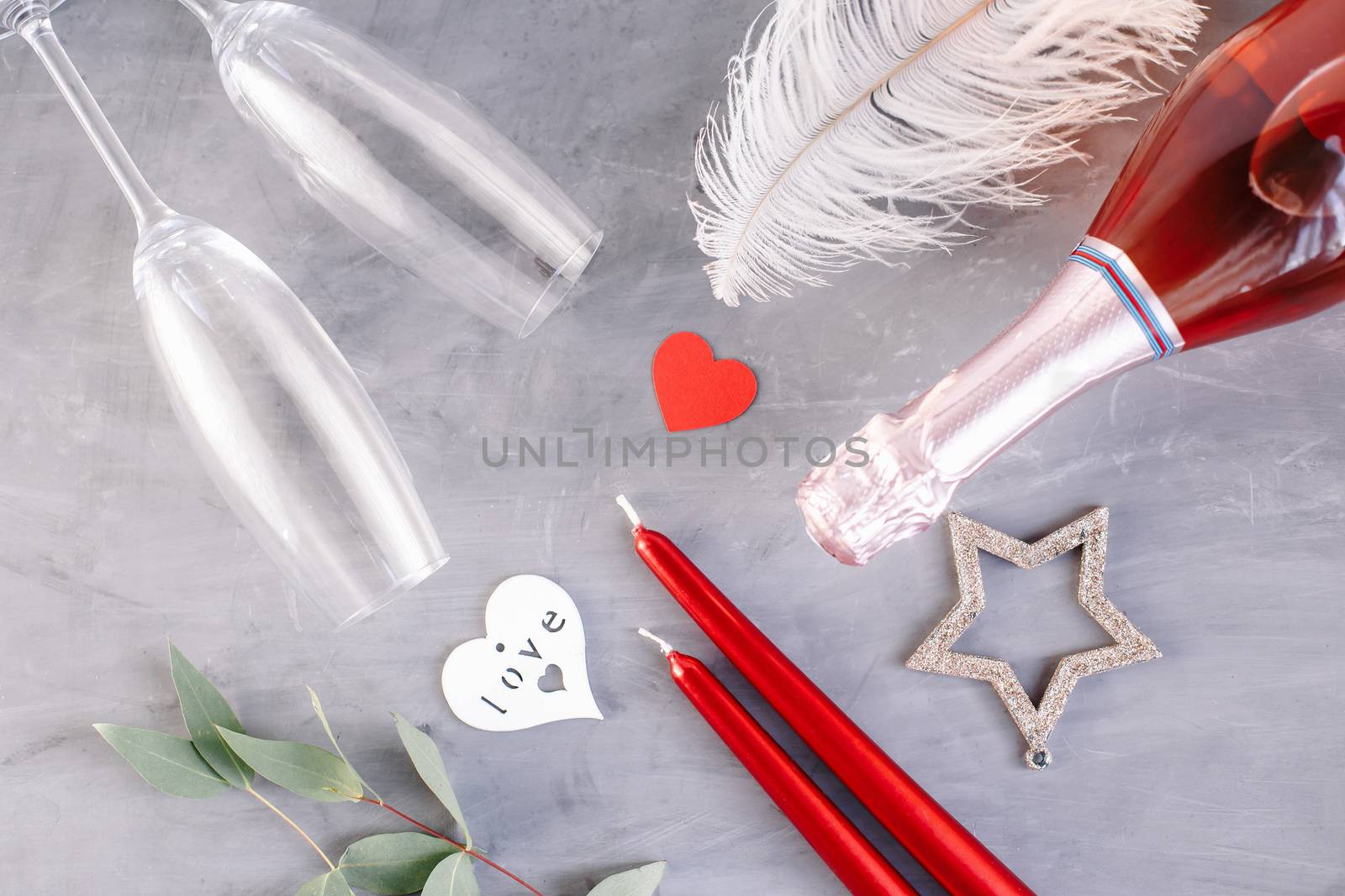 Flat lay composition with bottle of champagne, candles, feather and hearts for celebration on grey concrete background. Glass for champagne