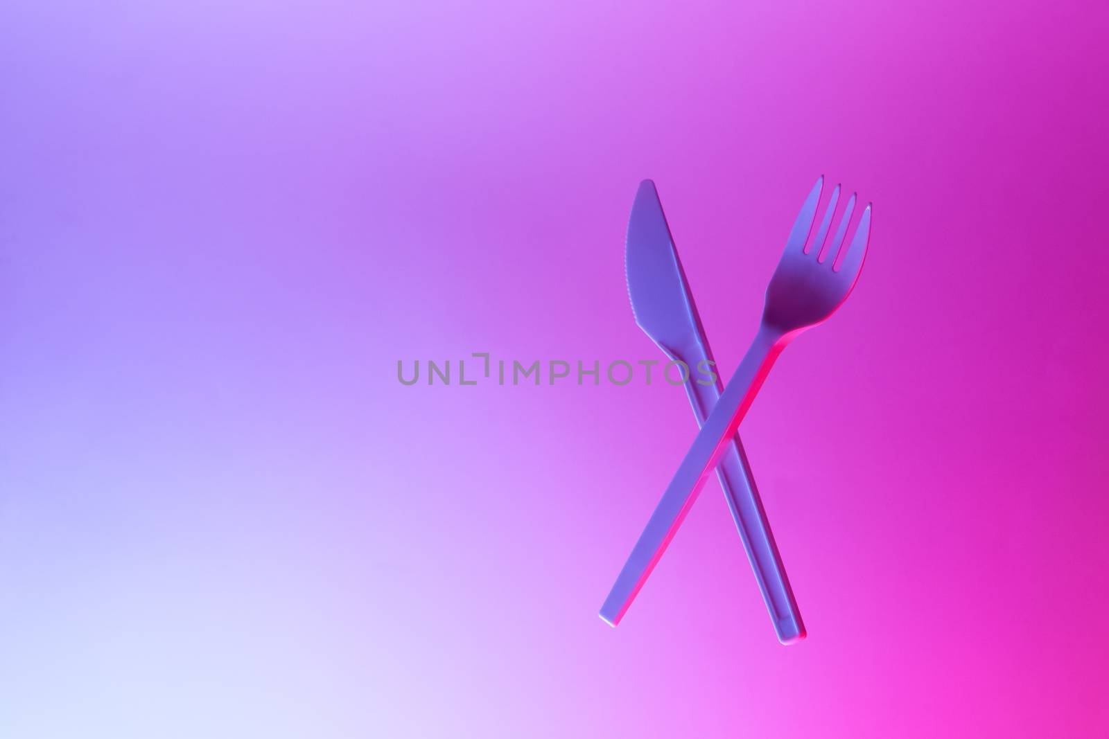 Disposable appliances plastic white fork and knife levitate in neon light, pink blue background, copy space. Eco, zero waste concept. Front view. Horizontal.