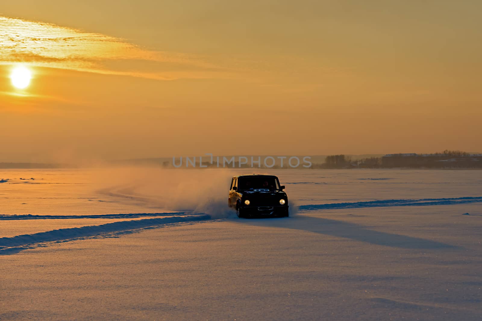The car rides on the ice of a frozen river in the snow in the rays of the setting sun.