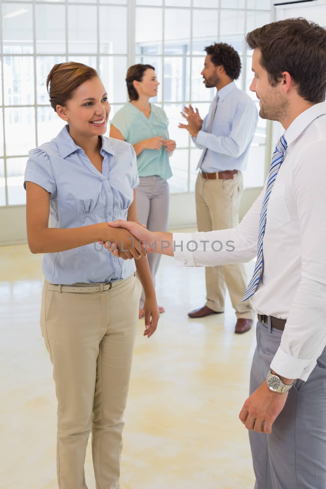 Two smiling young business people shaking hands with colleagues in background at the office