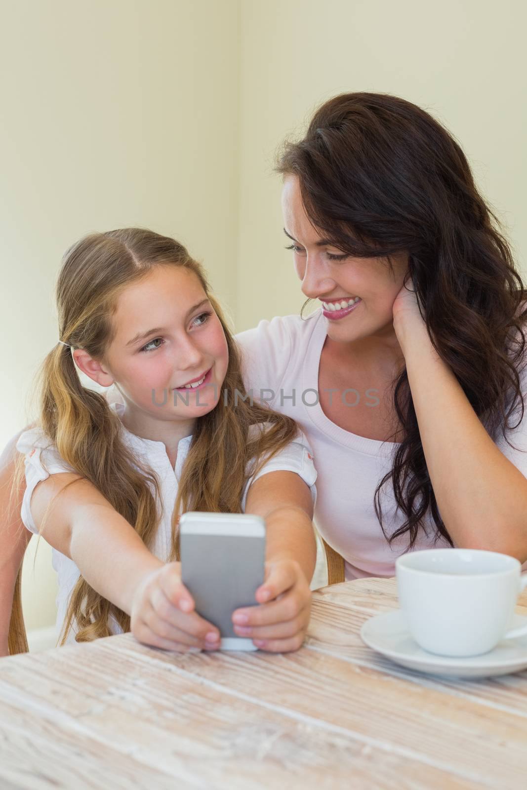 Cute little girl with mother photographing through mobile phone at table in house