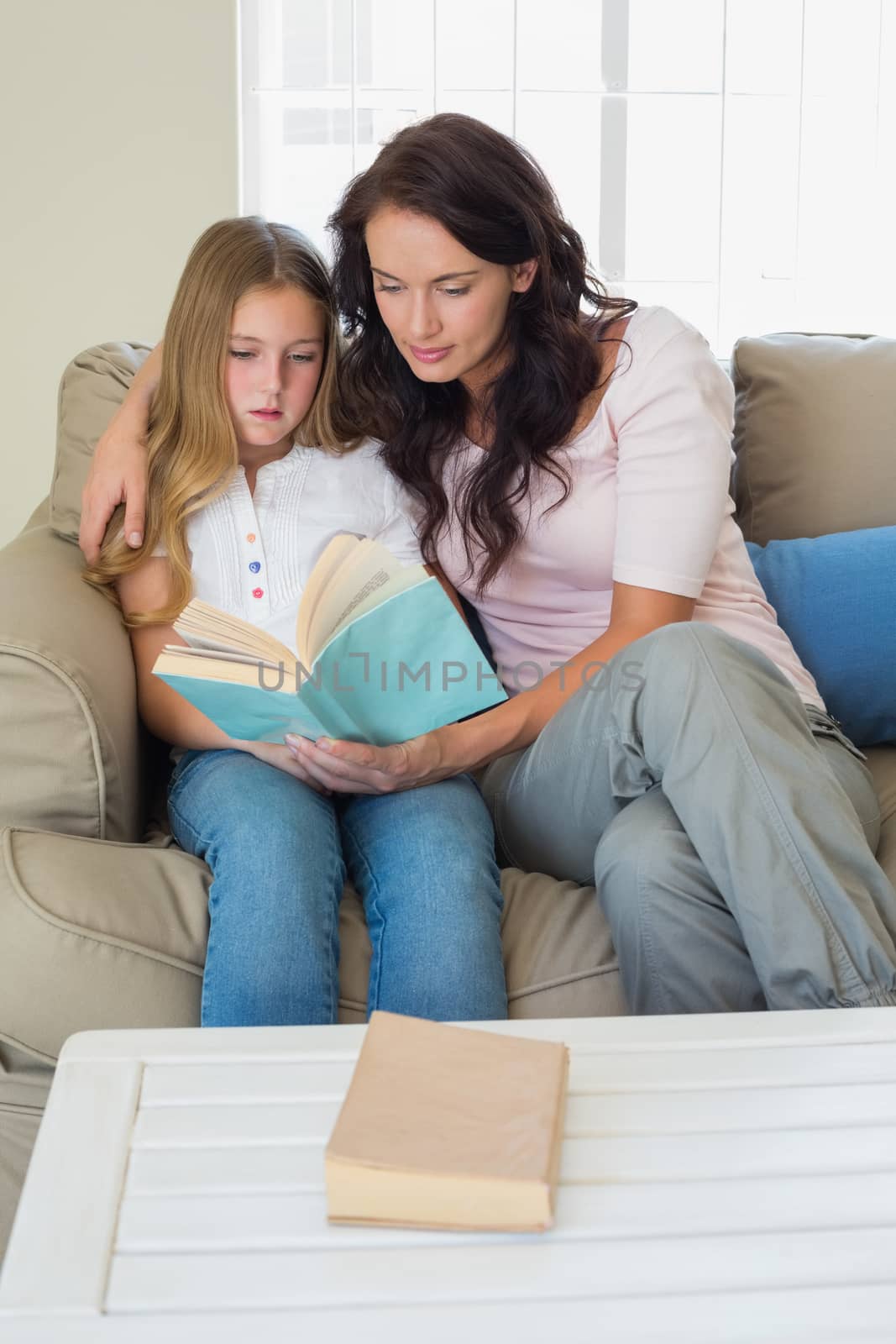 Little girl and mother reading novel together on sofa at home