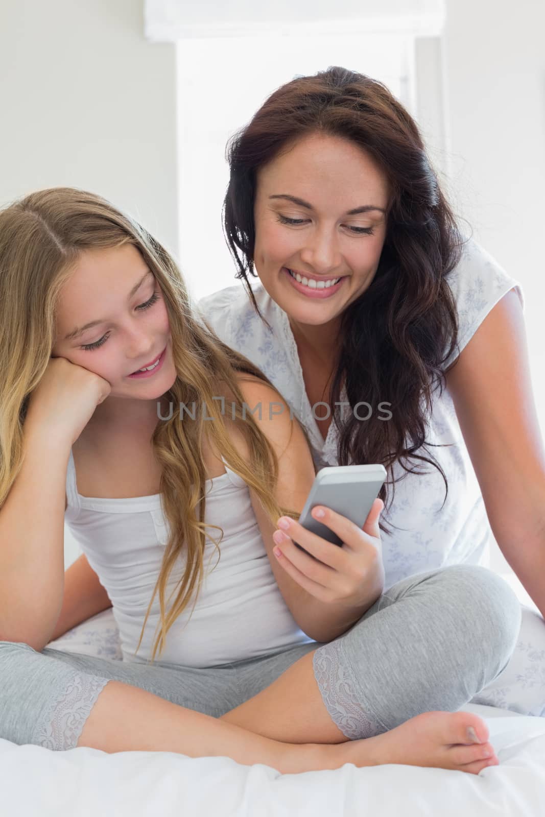 Mother and daughter using mobile phone together while sitting in bed at home