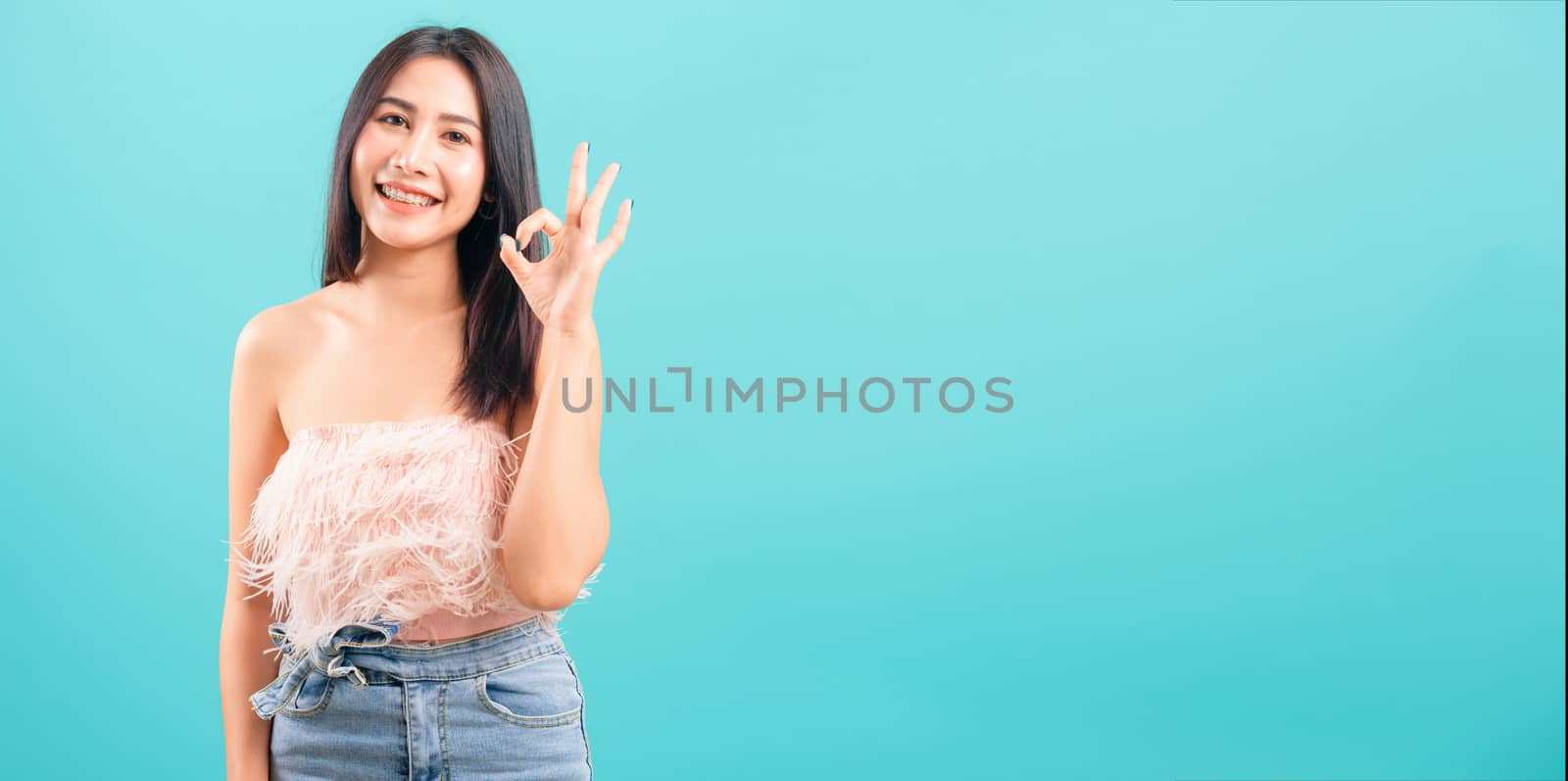 Smiling face asian beautiful woman her showing OK hand sign and looking to camera on blue background, with copy space for text