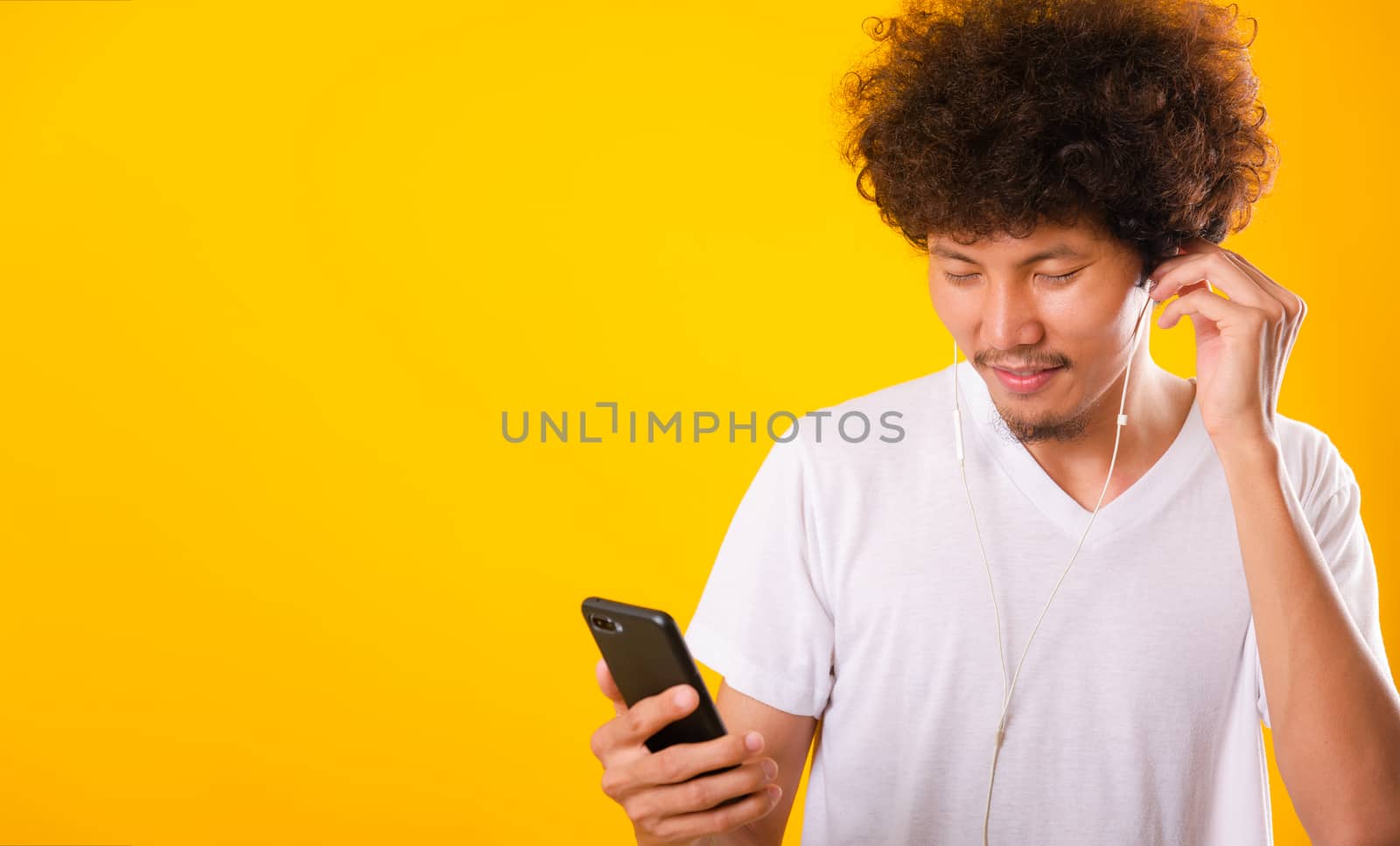 Happy asian handsome man with curly hair he smiling enjoying listening to music on earphones using a mobile smartphone isolate on yellow background