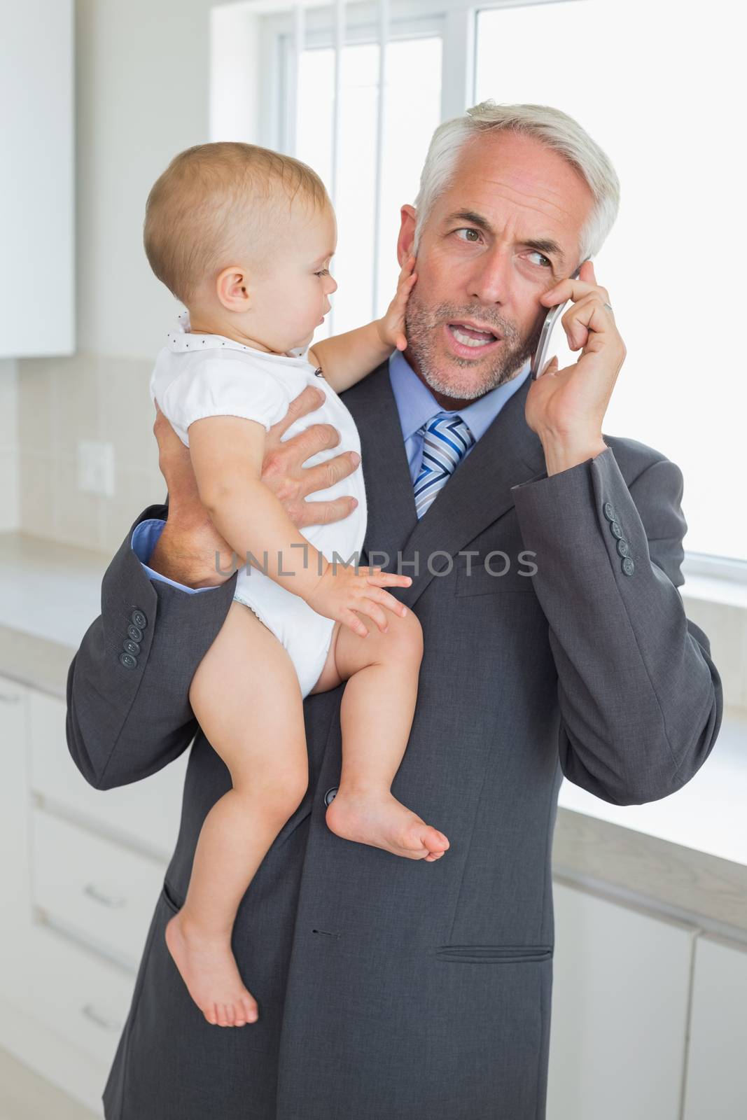 Distracted businessman holding his baby in the morning before work at home in the kitchen