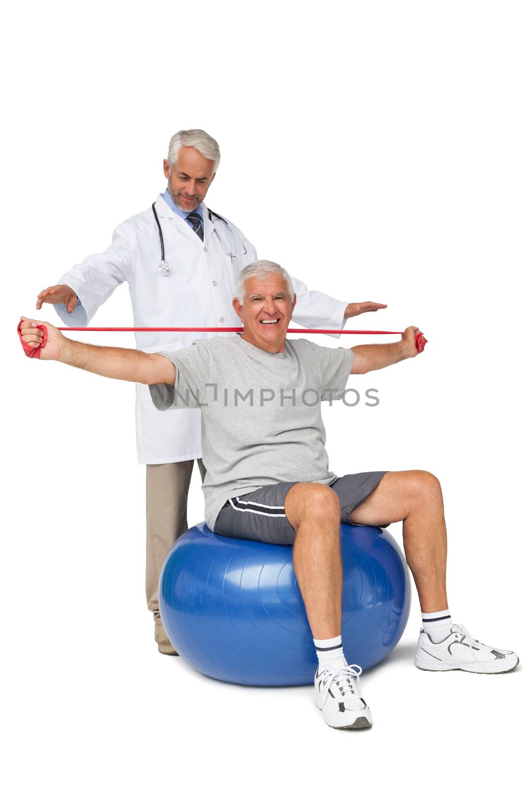 Male physiotherapist looking at senior man sit on exercise ball with yoga belt over white background