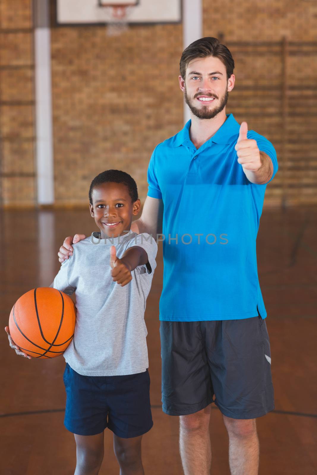 Portrait of sports teacher and schoolboy showing thumbs up in basketball court at school gym