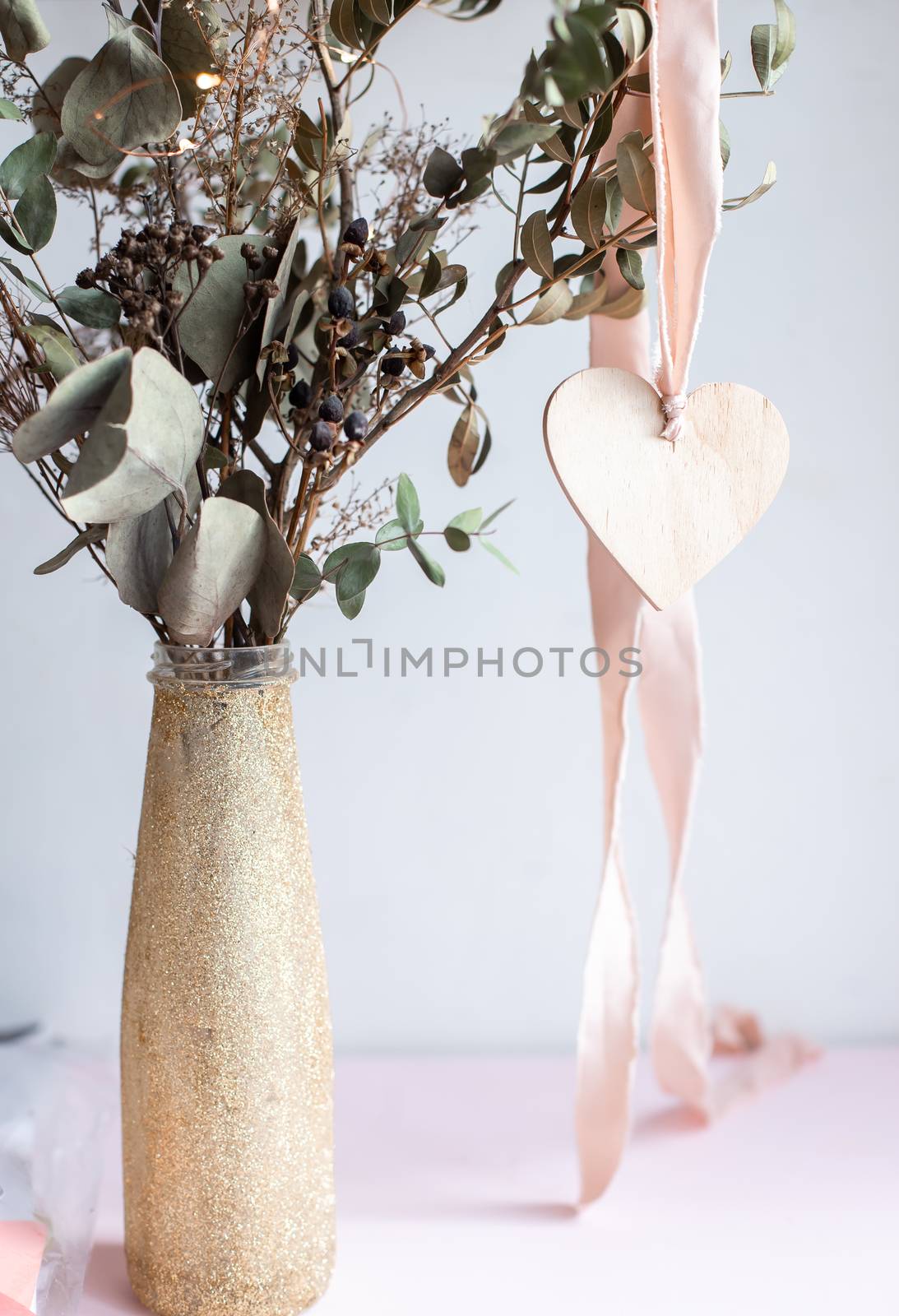 Light wooden heart pendant with pink silk ribbon on a background of a golden shiny glittered vase with dried eucalyptus branches.