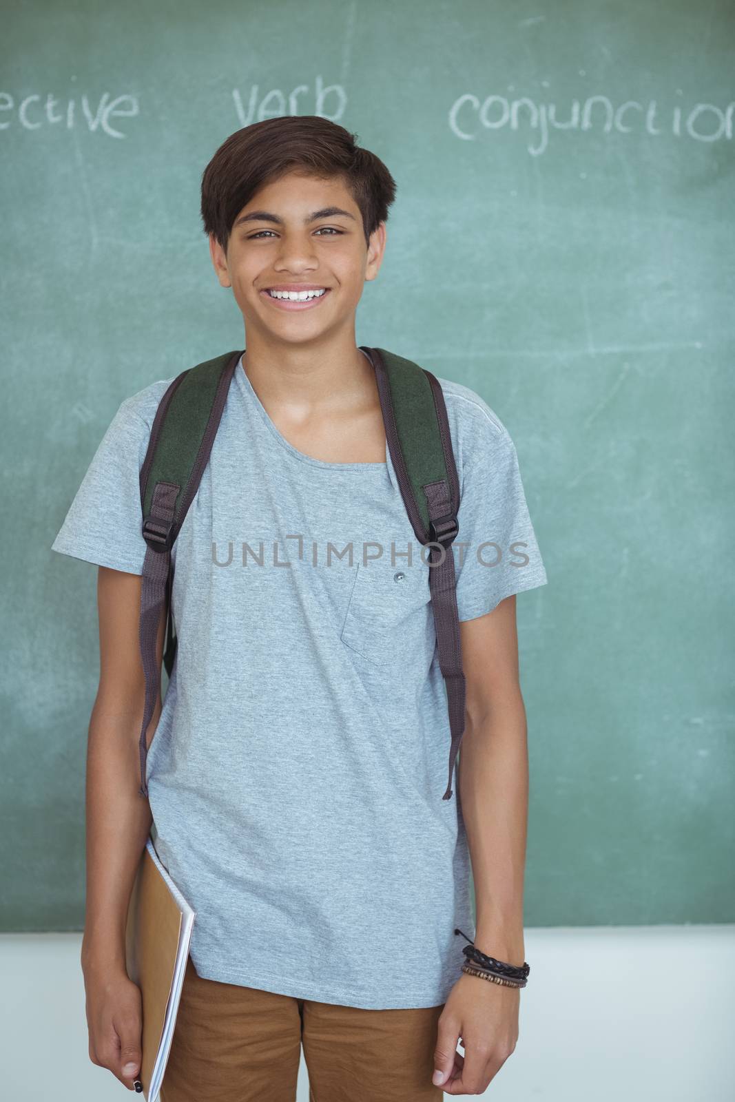 Smiling schoolboy with backpack and book standing against chalkboard in classroom at school