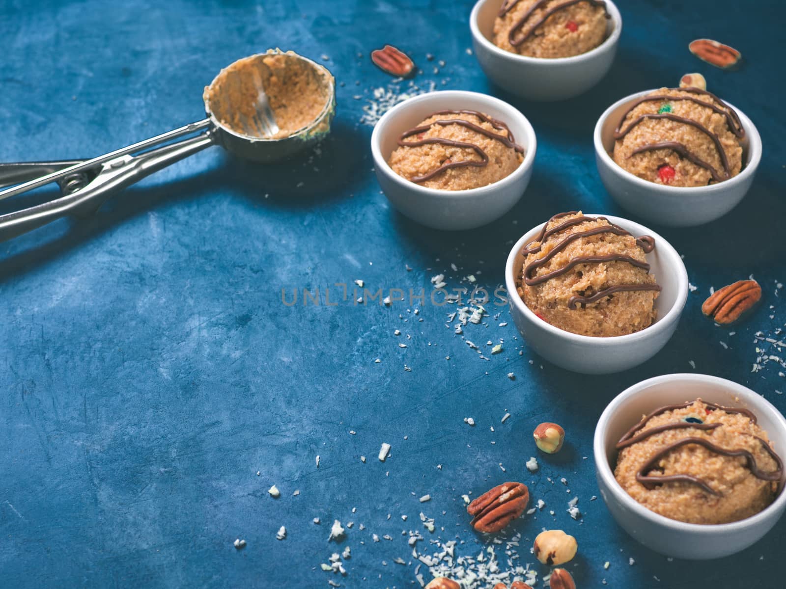 Safe-to-eat raw monster cookie dough in small portion bowl, ice cream scoop and nuts on blue background. Ideas and recipes for kids and toddlers meal. Copy space for text.