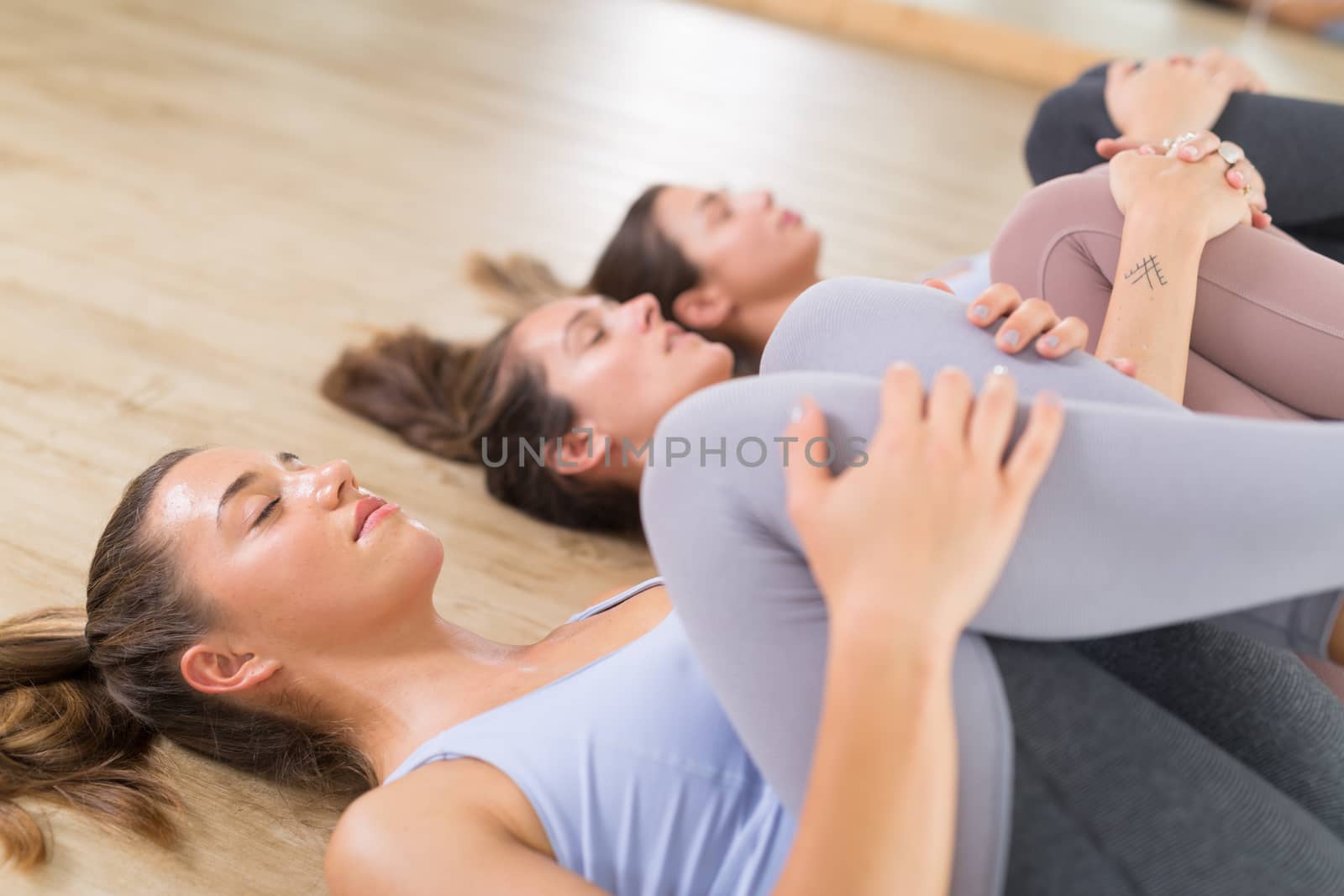 Group of three young sporty attractive women in yoga studio, lying on the floor, stretching and relaxing after the workout. Healthy active lifestyle, working out indoors in gym.