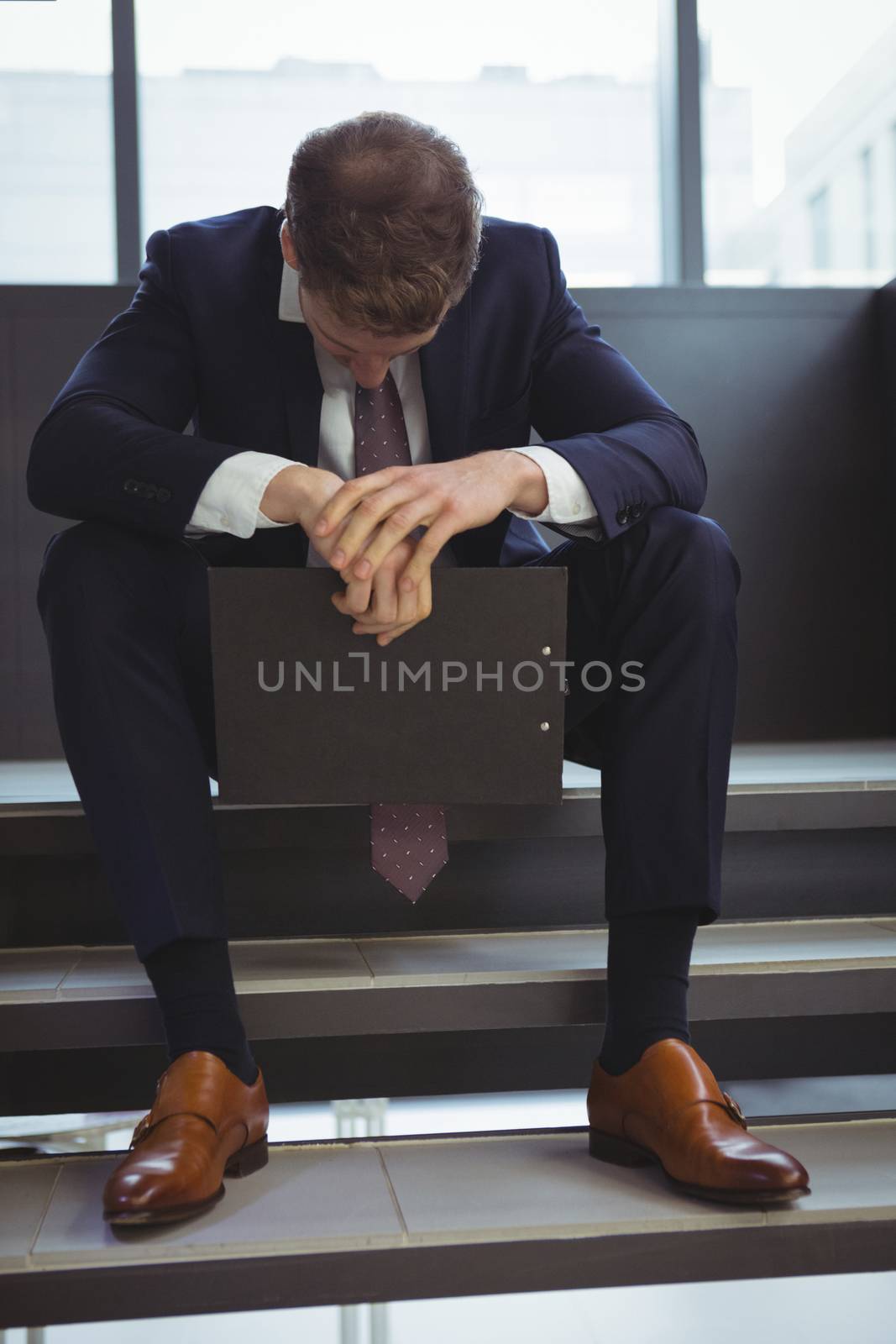 Depressed businessman with clipboard sitting on stairs at office