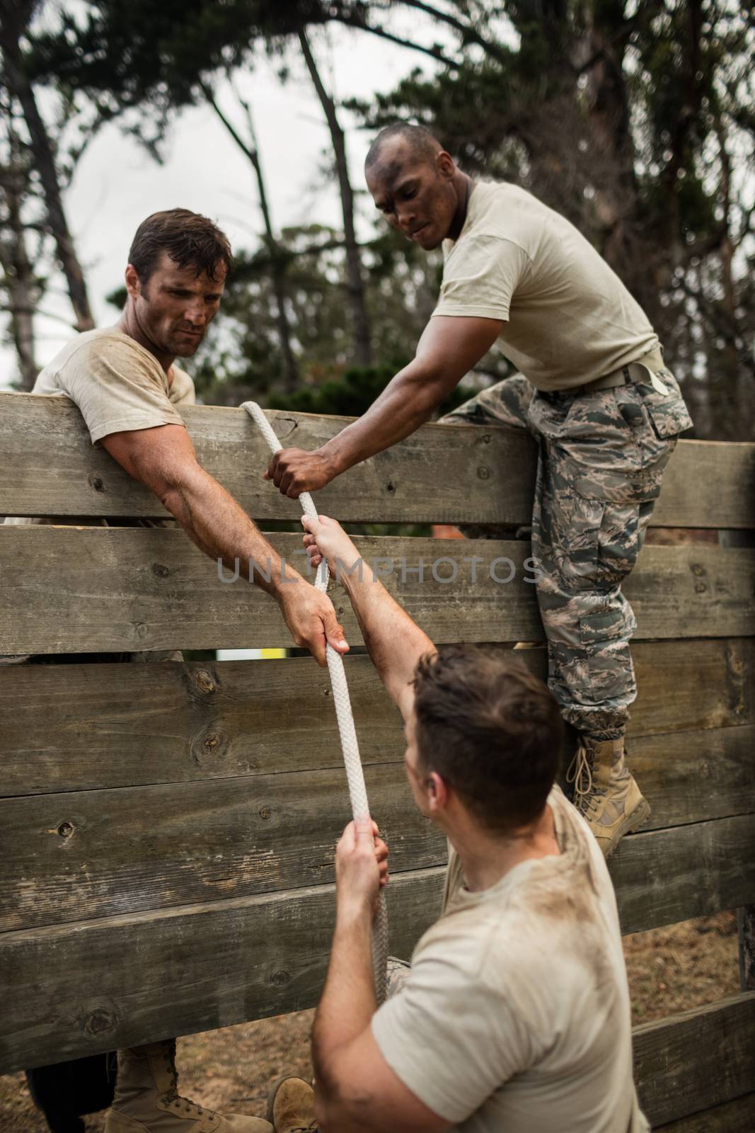 Soldiers climbing wooden wall with rope in boot camp