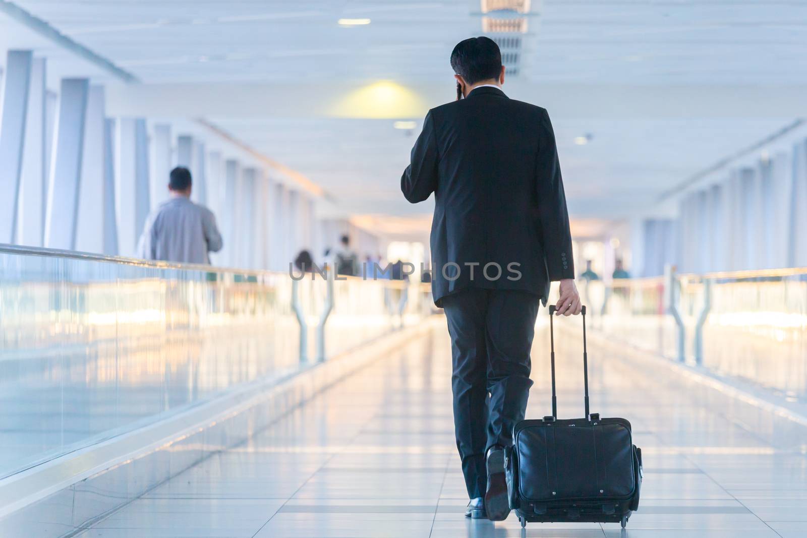 Rear view of unrecognizable formaly dressed businessman walking and wheeling a trolley suitcase at the lobby, talking on a mobile phone. Business travel concept.