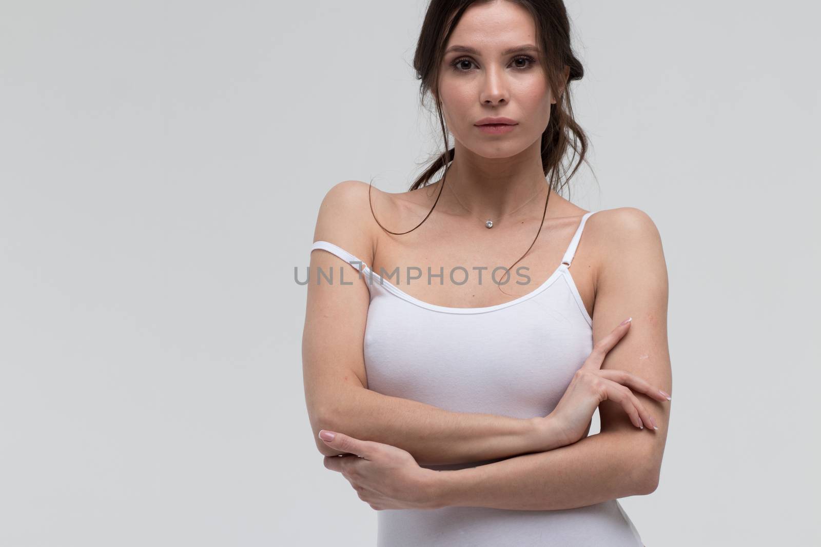 Relaxed sensual brunette woman in white t shirt pulling thin strap while looking at camera on white background