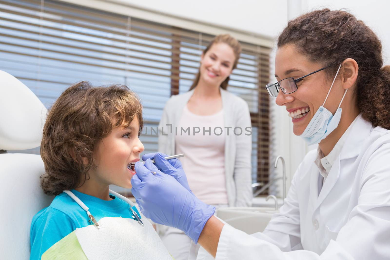 Pediatric dentist examining a little boys teeth with his mother watching at the dental clinic